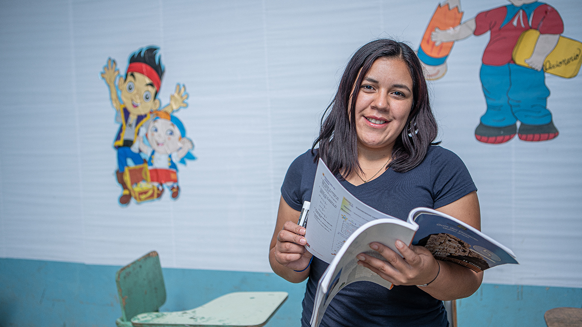Une femme se tient debout dans une salle de classe, tournant la page d’un livre en souriant à l’objectif.